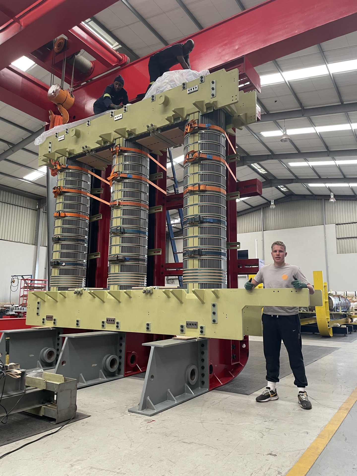 Matteo Buxman standing beside a fully assembled three-limb transformer core and coil assembly, with technicians working on the top yoke